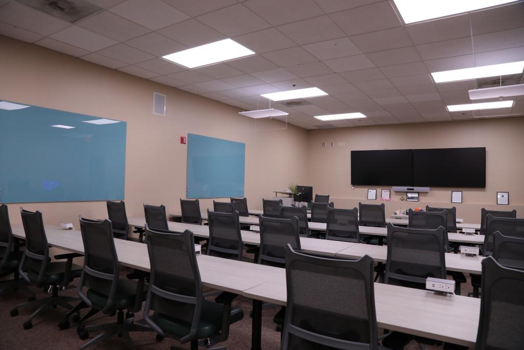 Two TVs are mounted on the far wall with rows of tables and chairs facing them. Two blue squares are on the right wall.